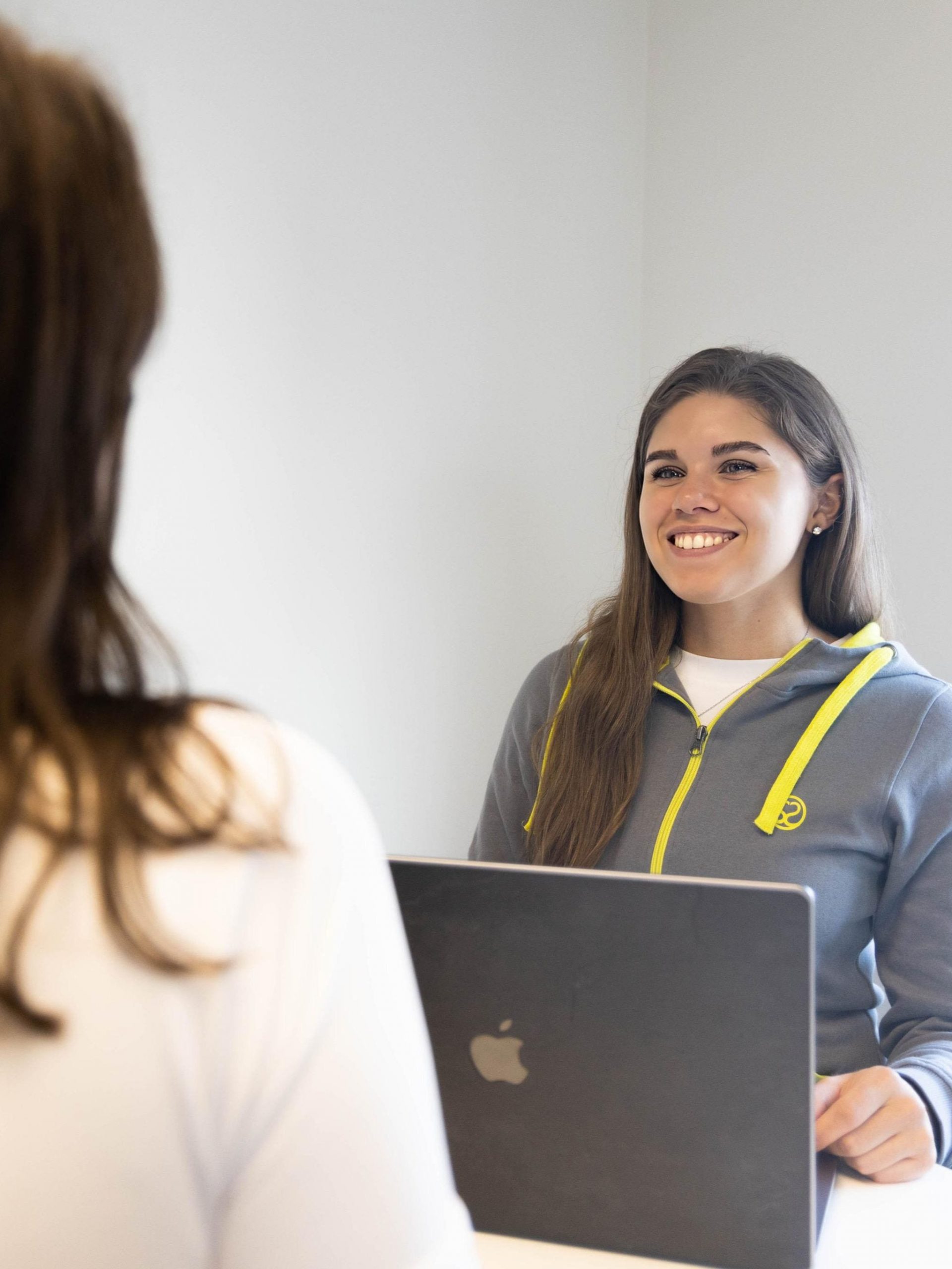 Eine freundlich lächelnde Fitnesstrainerin siitzt vor einem Laptop und berät eine Kundin