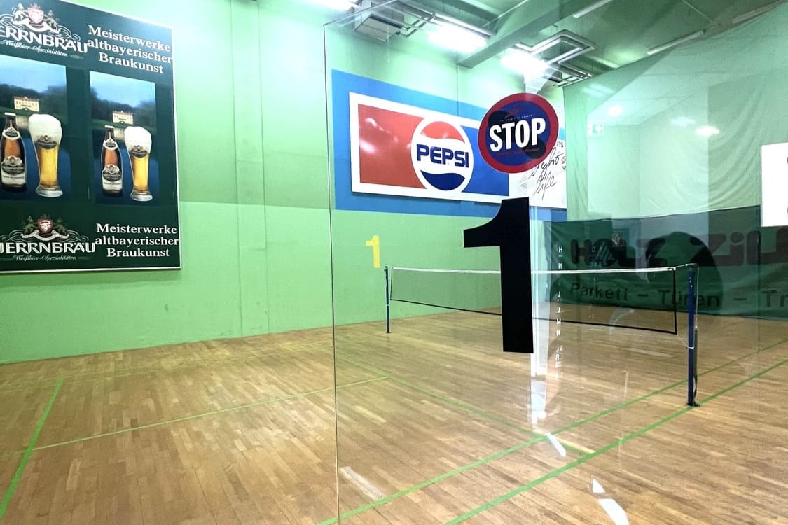 Badminton Court 1 hinter einer Glastür mit gespanntem Badminton-Netz und Werbung an der Wand