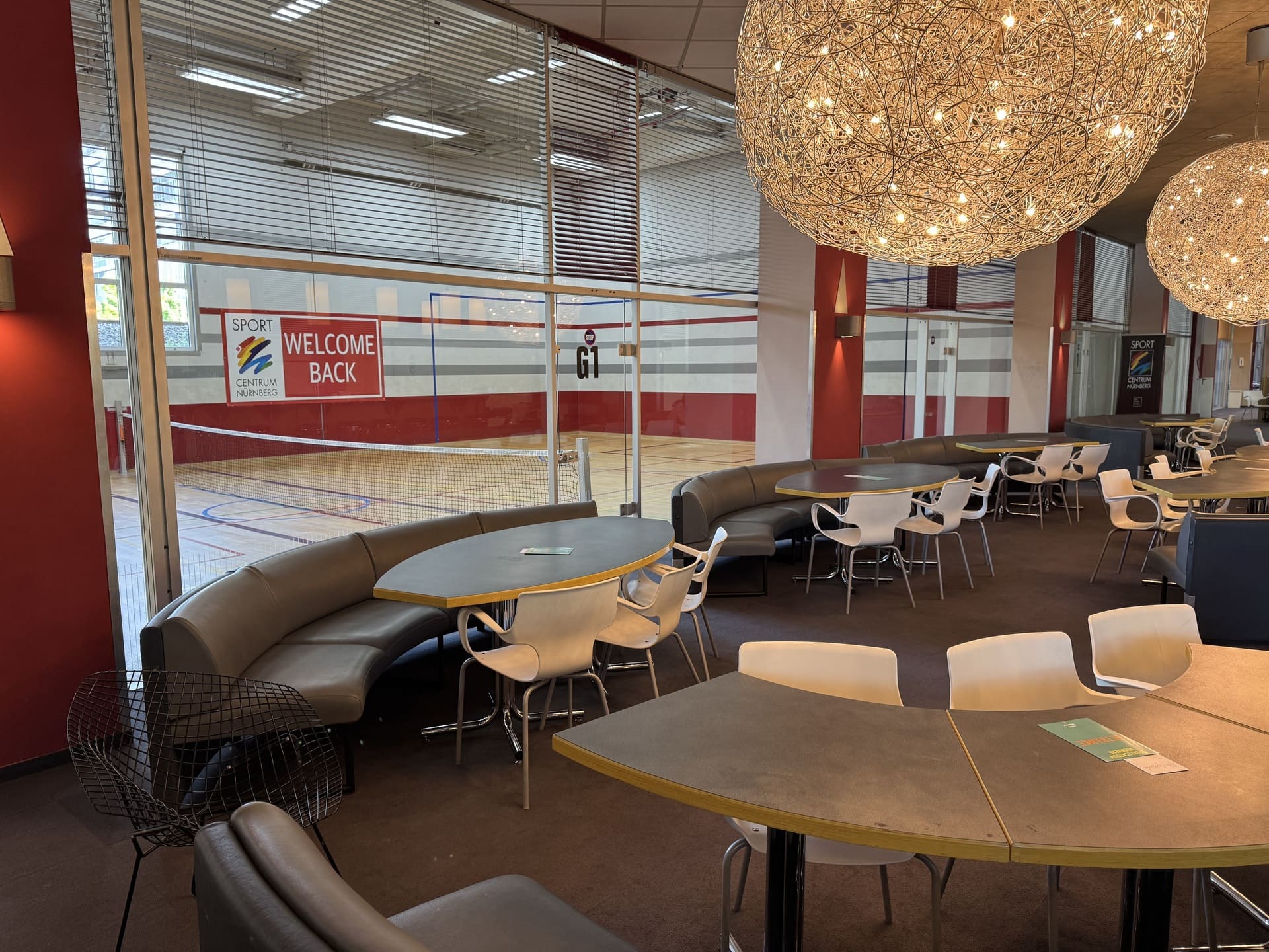 Lounge-Bereich mit Stühlen, Bänken und Tischen sowie Blick durch eine Glaswand in den Game Court im Sportcentrum Nürnberg