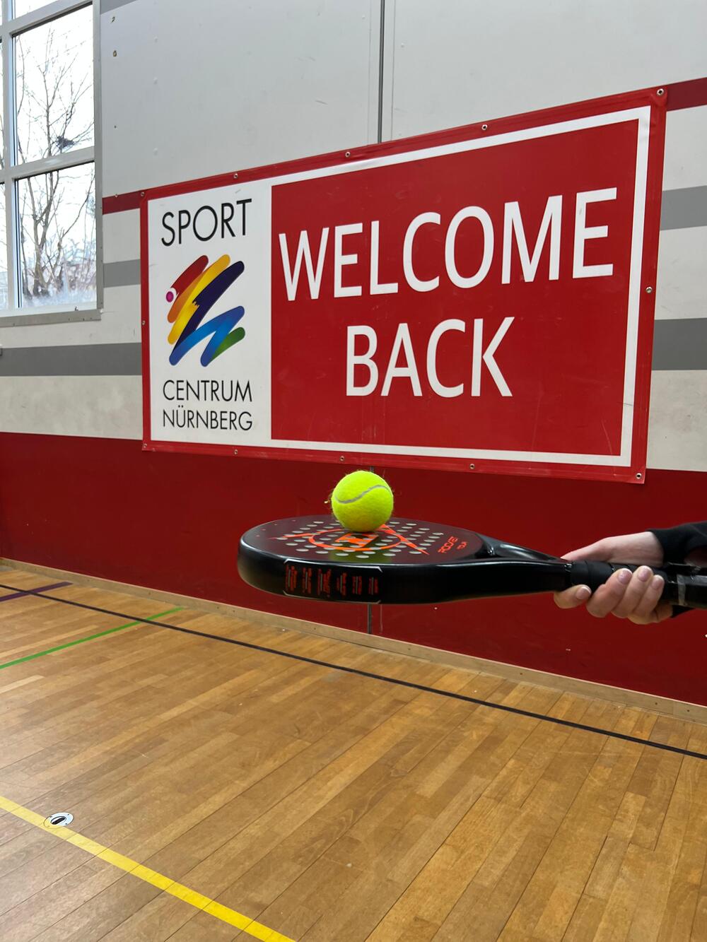 Tennisball liegt auf einem Padel Tennis-Schläger vor dem Tennisnetz im Game Court auf dessen Wand "Welcome Back" steht.