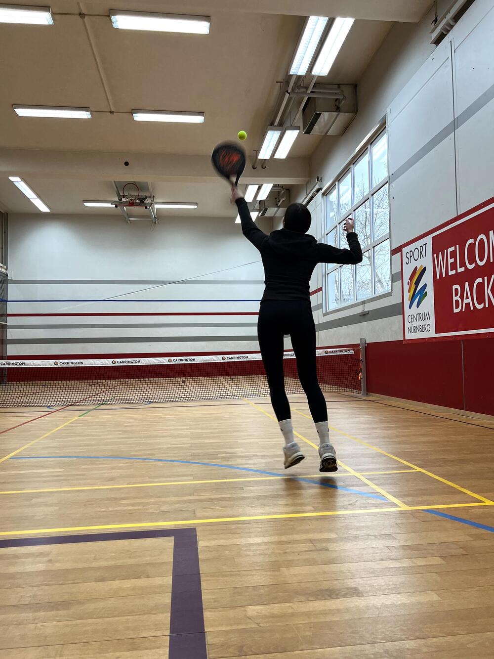 Junge Frau springt, um mit dem Padel Tennis-Schläger den ankommenden Tennisball zu erwischen im Game Court des Sportcentrum Nürnberg