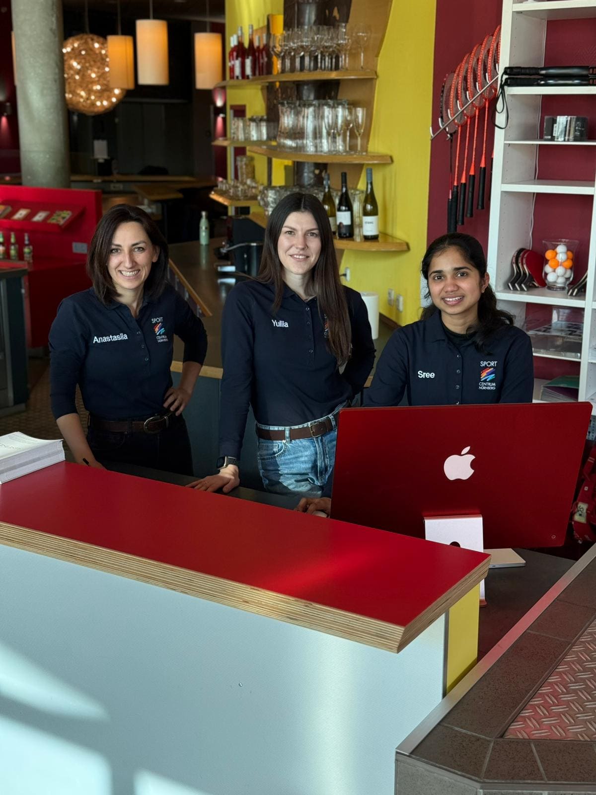 Drei junge Frauen stehen gemeinsam hinter der Empfangstheke des Sportcentrum Nürnbergs und lächeln in die Kamera